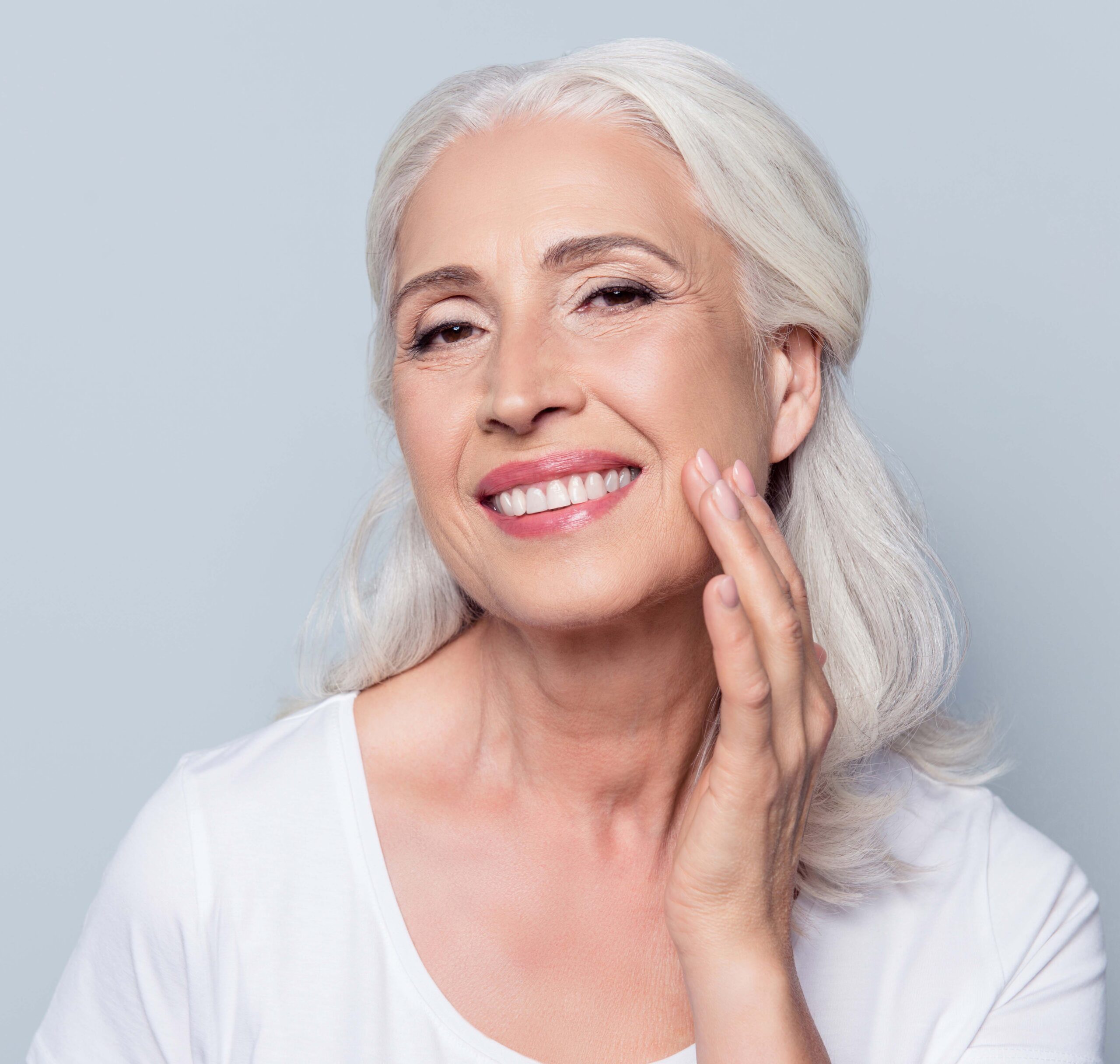 Charming, pretty, old woman touching her perfect soft face skin with fingers, smiling at camera over gray background, using day, night face cream, cosmetology procedures Juvéderm® / Other Fillers in Savannah, GA